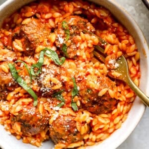 Overhead shot of meatball parmesan orzo in a bowl, topped with basil and parmesan, with a fork resting in it.