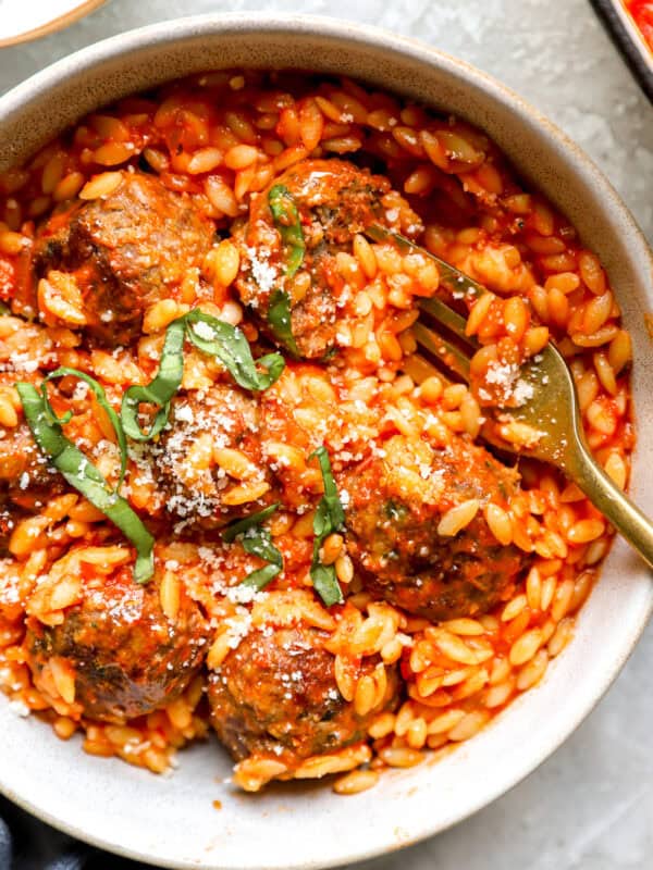 Overhead shot of meatball parmesan orzo in a bowl, topped with basil and parmesan, with a fork resting in it.