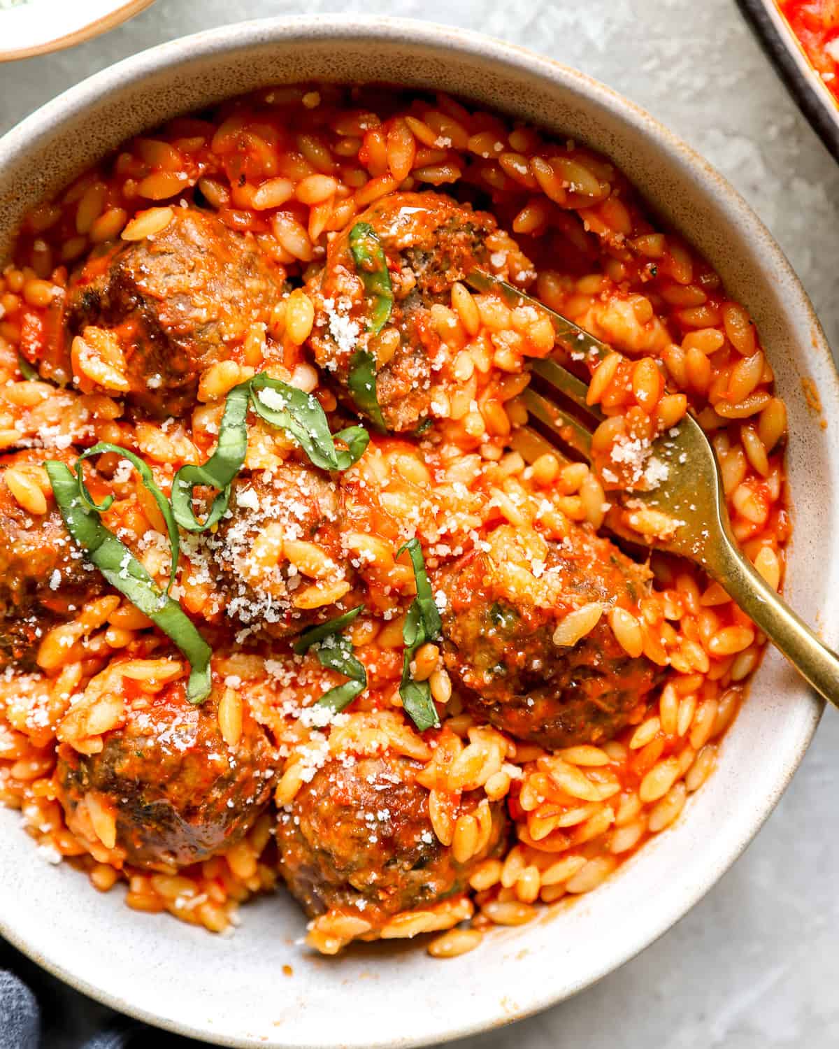 Overhead shot of meatball parmesan orzo in a bowl, topped with basil and parmesan, with a fork resting in it.