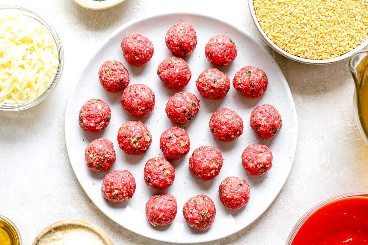 Rolled raw meatballs arranged on a white plate, ready to be browned for meatball parmesan orzo.