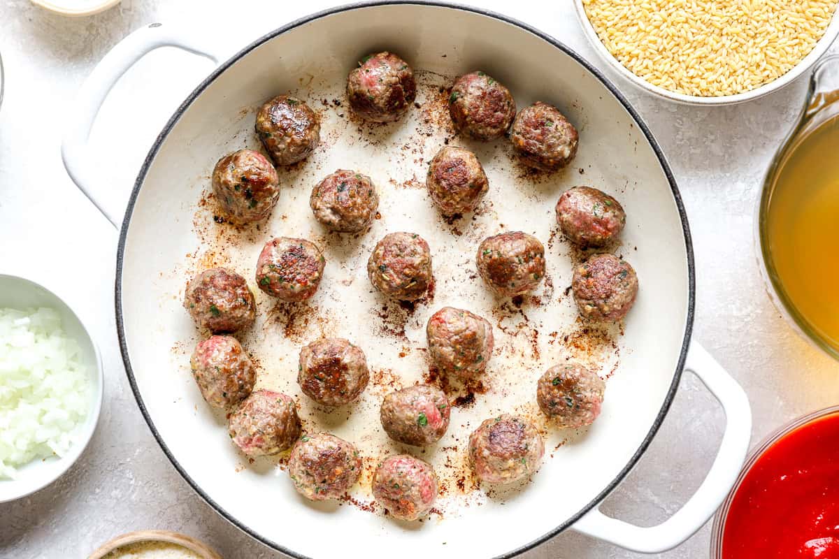 Browned meatballs in a white skillet, seared and ready for the tomato orzo sauce.