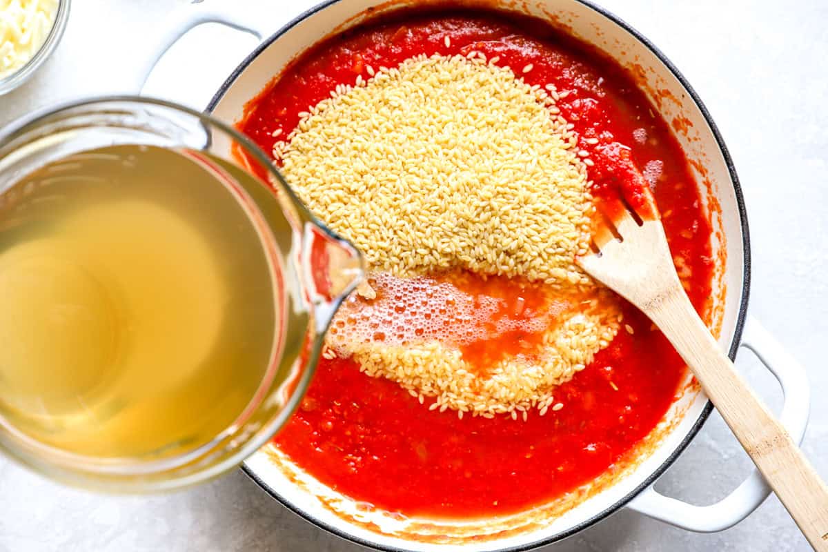 Dry orzo and chicken broth being added to tomato sauce in a skillet for meatball parmesan orzo.