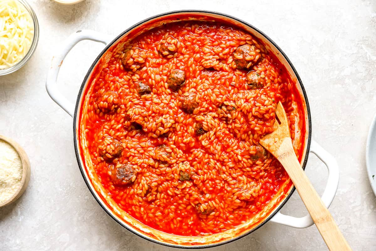 Orzo simmered in tomato sauce with meatballs nestled throughout, stirred with a wooden spoon.
