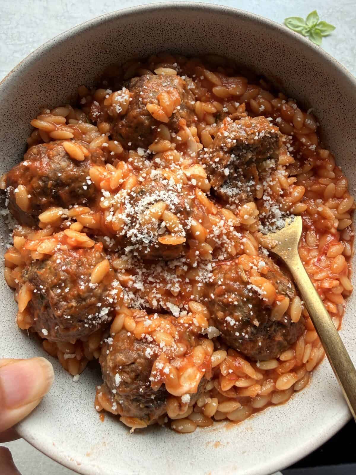 Overhead close-up of a bowl of meatball parmesan orzo, with a hand holding the bowl and a gold fork resting among the meatballs.