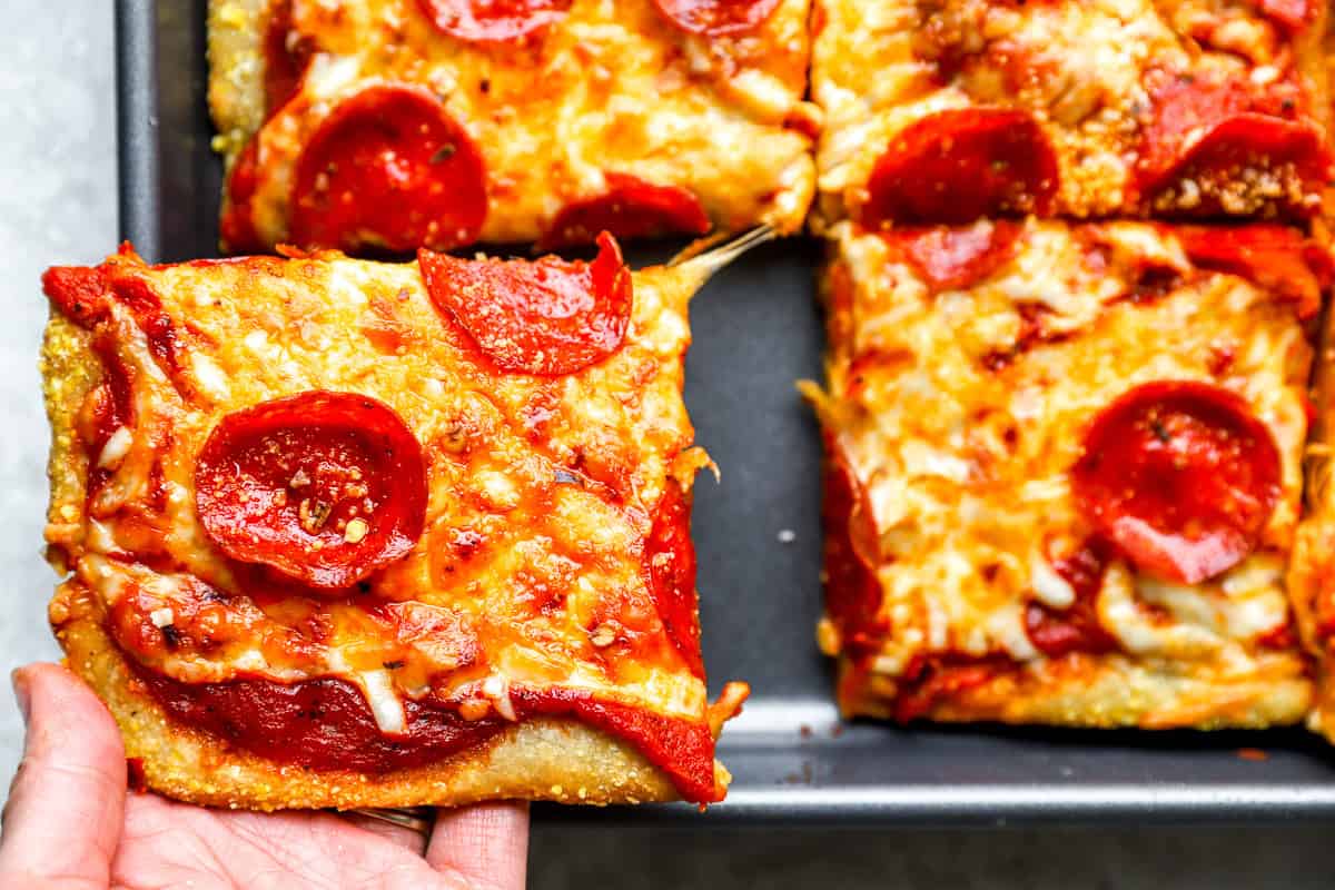 Hand lifting a cheesy pepperoni slice of sheet pan pizza with a stretchy cheese pull