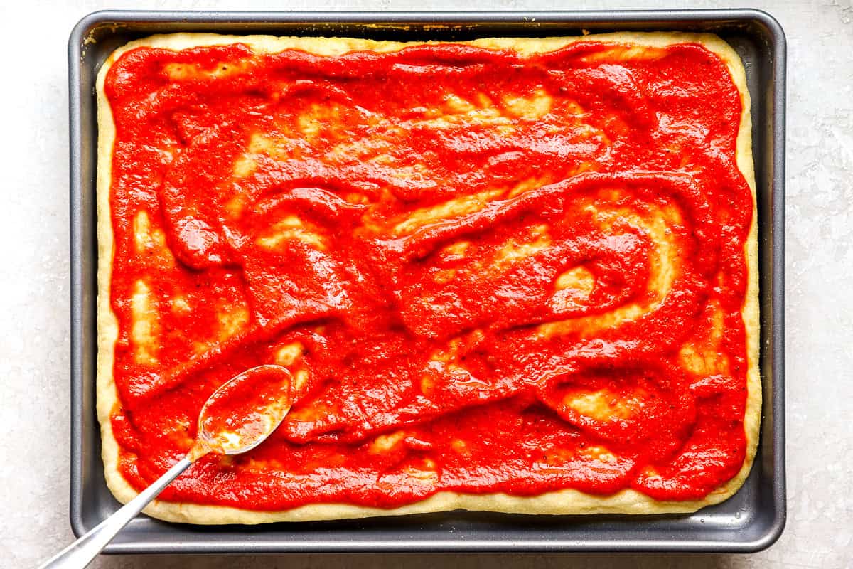 Pizza sauce being spread over the par-baked crust with the back of a spoon