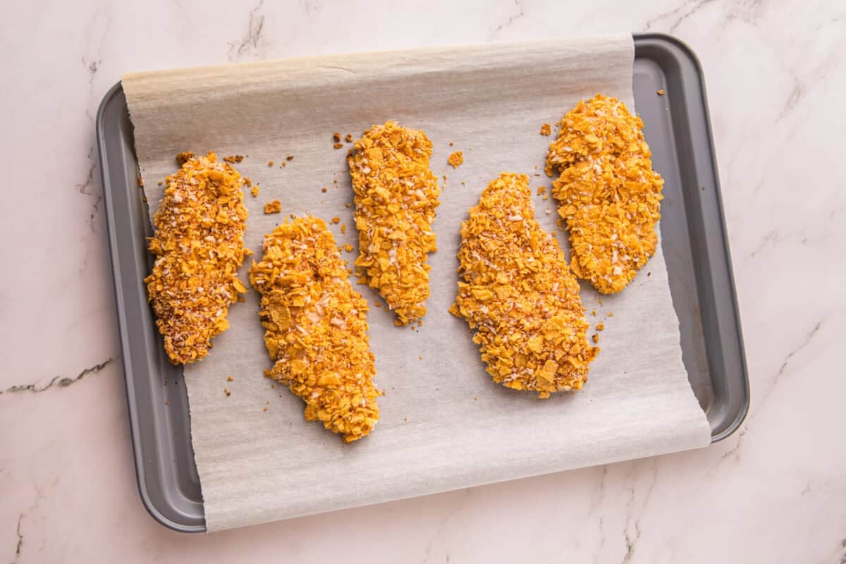 Five golden brown cornflake chicken tenders on a baking sheet after baking