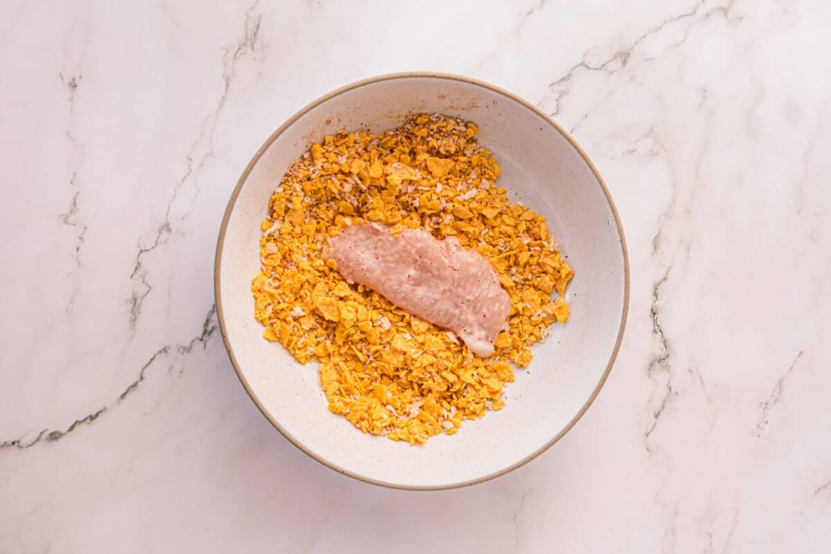 Raw chicken tenderloin resting on the cornflake mixture before being pressed and coated