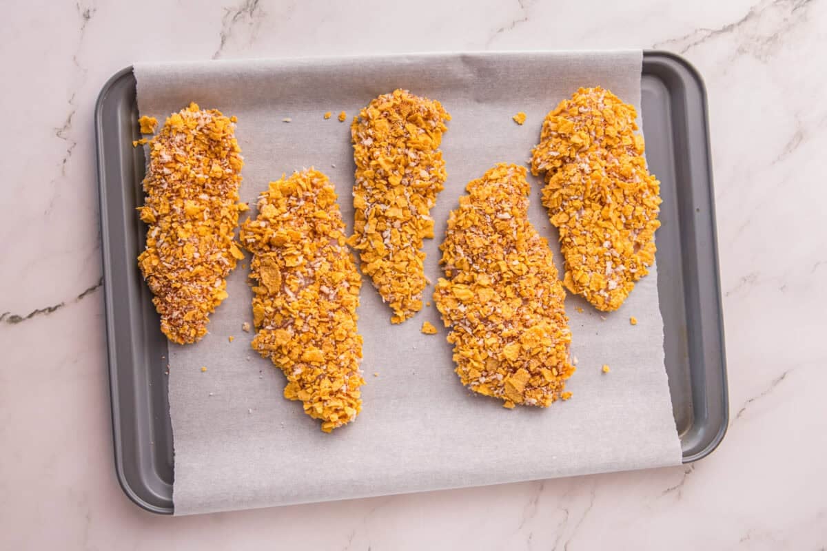 Five cornflake-coated chicken tenders arranged on a parchment-lined baking sheet before baking