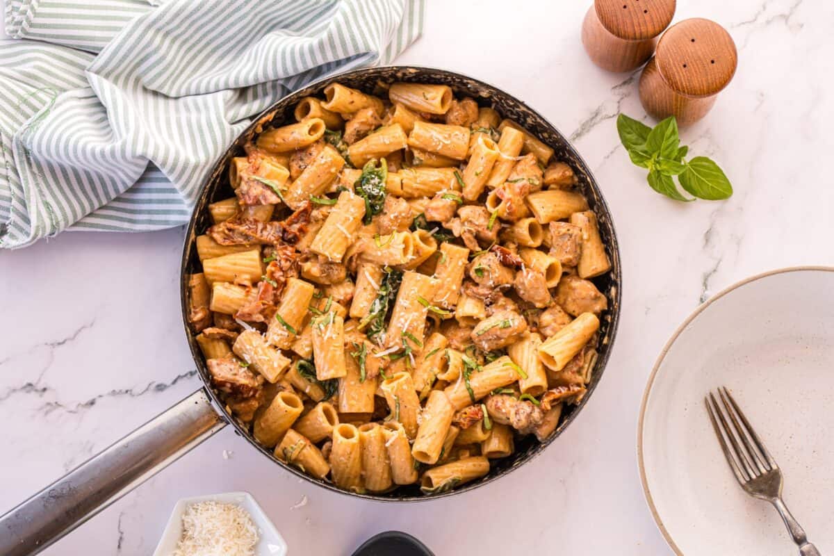 Finished creamy Tuscan chicken pasta in a skillet, garnished with basil and Parmesan and served with a striped napkin.