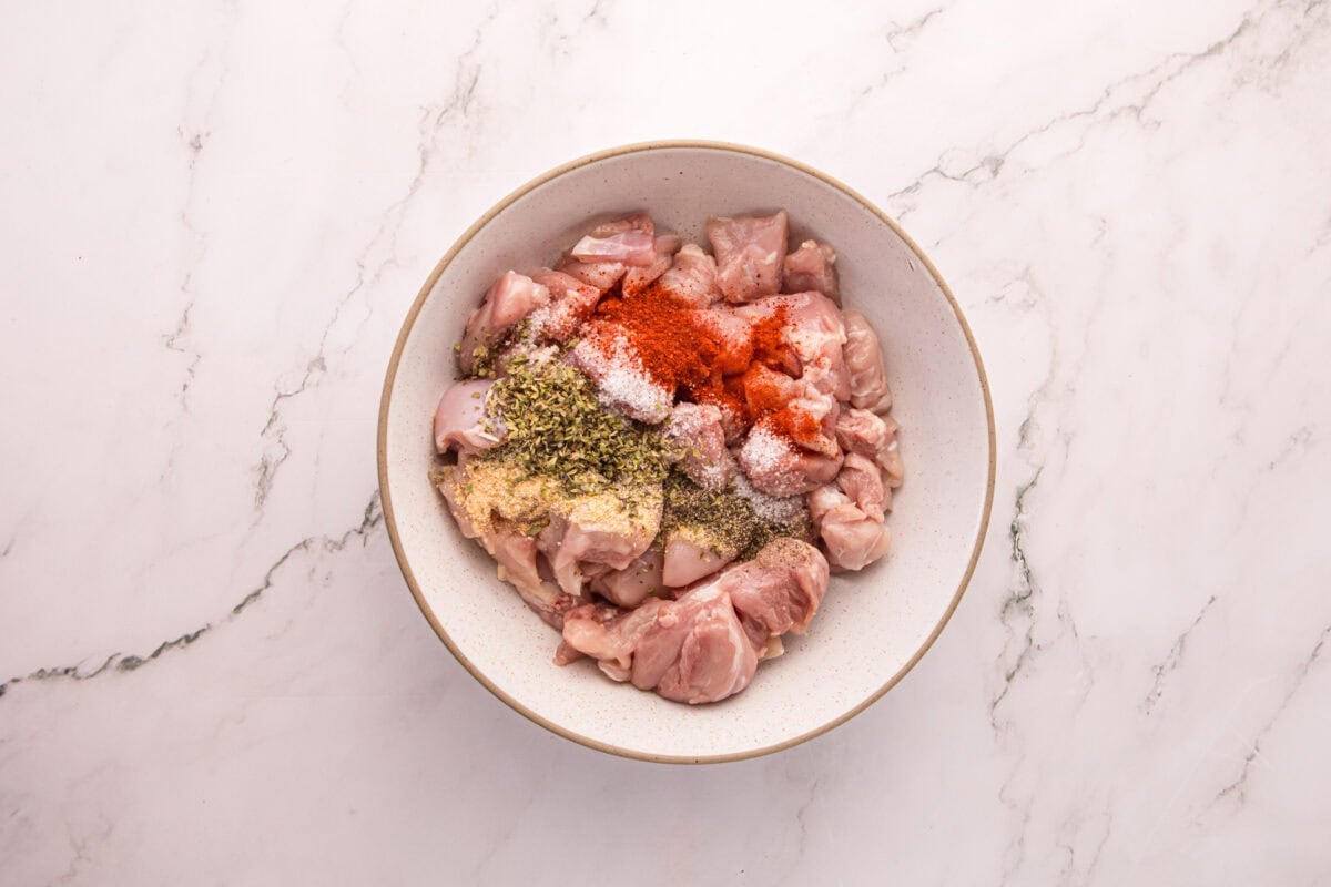 Raw chicken pieces in a white bowl topped with salt, smoked paprika, garlic powder, black pepper, and Italian seasoning.