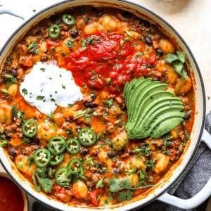 Overhead shot of taco gnocchi skillet topped with salsa, sour cream, sliced avocado, jalapeños, and cilantro in a white Dutch oven.