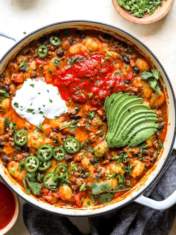 Overhead shot of taco gnocchi skillet topped with salsa, sour cream, sliced avocado, jalapeños, and cilantro in a white Dutch oven.