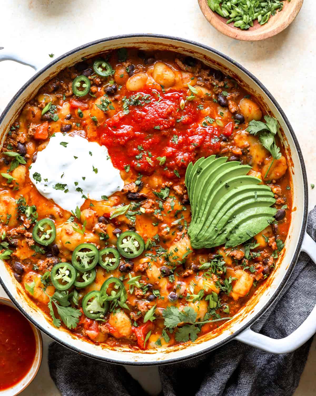 Overhead shot of taco gnocchi skillet topped with salsa, sour cream, sliced avocado, jalapeños, and cilantro in a white Dutch oven.